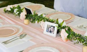 Wedding reception table decorated with pink tablecloth, greenery garland, white roses and a small framed photo centerpiece.