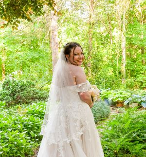 A bride in a flowing white lace dress walks through a sunlit forest setting surrounded by lush green foliage.