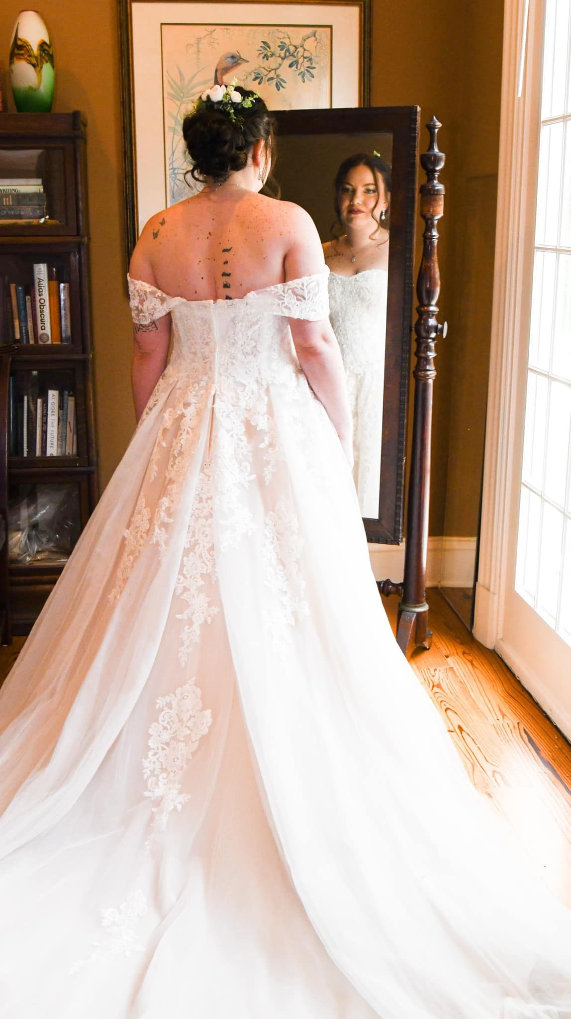 A wedding dress with off-shoulder design and lace details is shown from behind as someone looks in a mirror.