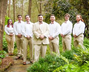 A wedding party in khaki and white attire poses on a stone path in a lush forest setting.