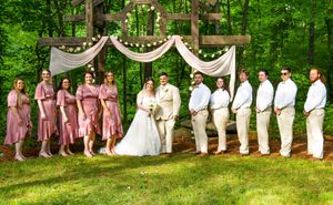 Wedding party lined up outdoors with bridesmaids in pink dresses and groomsmen in white under draped fabric decor.