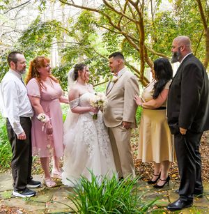 A wedding ceremony group gathers on a garden path surrounded by green foliage.