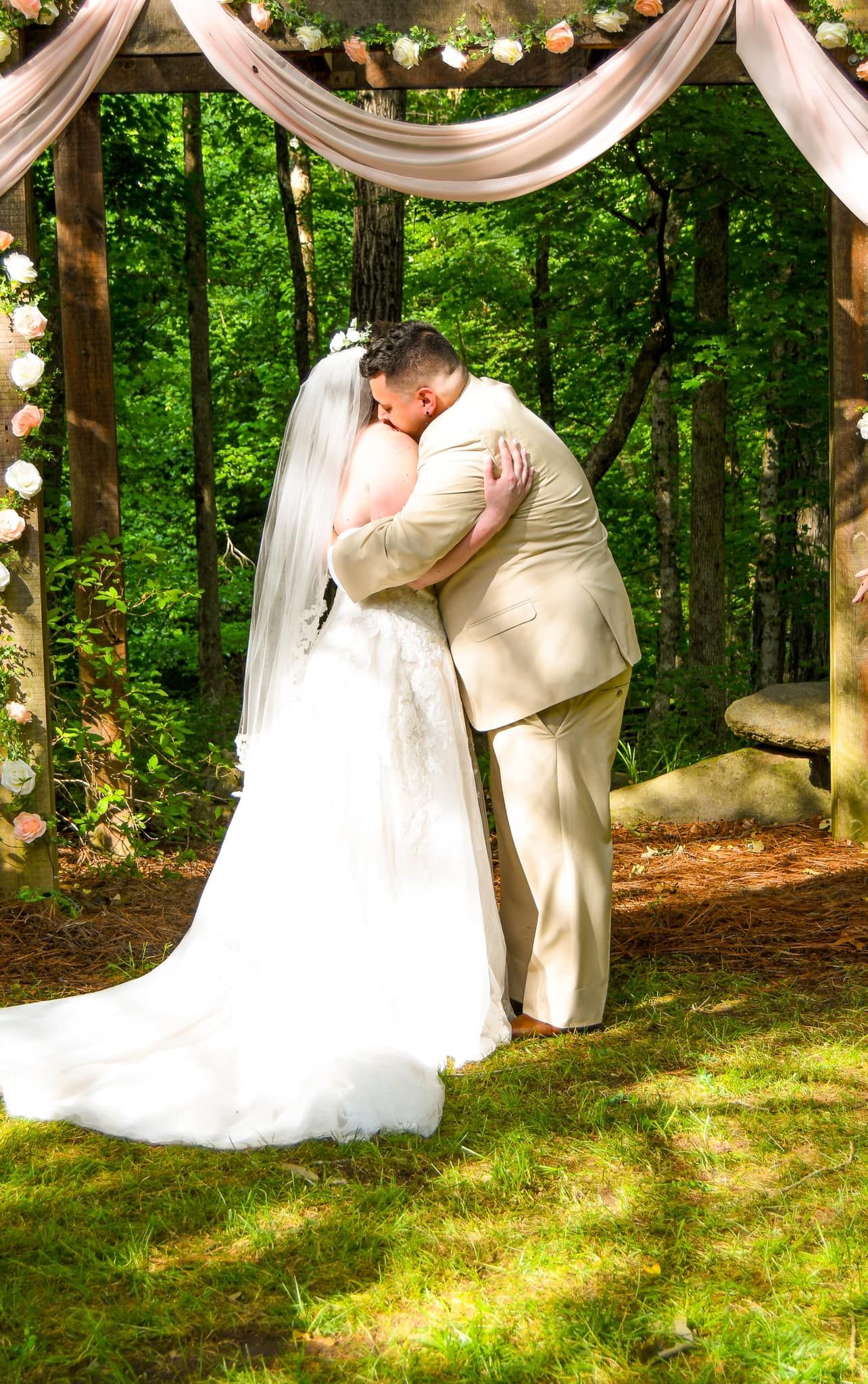 Wedding couple shares romantic kisses under a decorated outdoor arbor with white fabric draping and string lights.