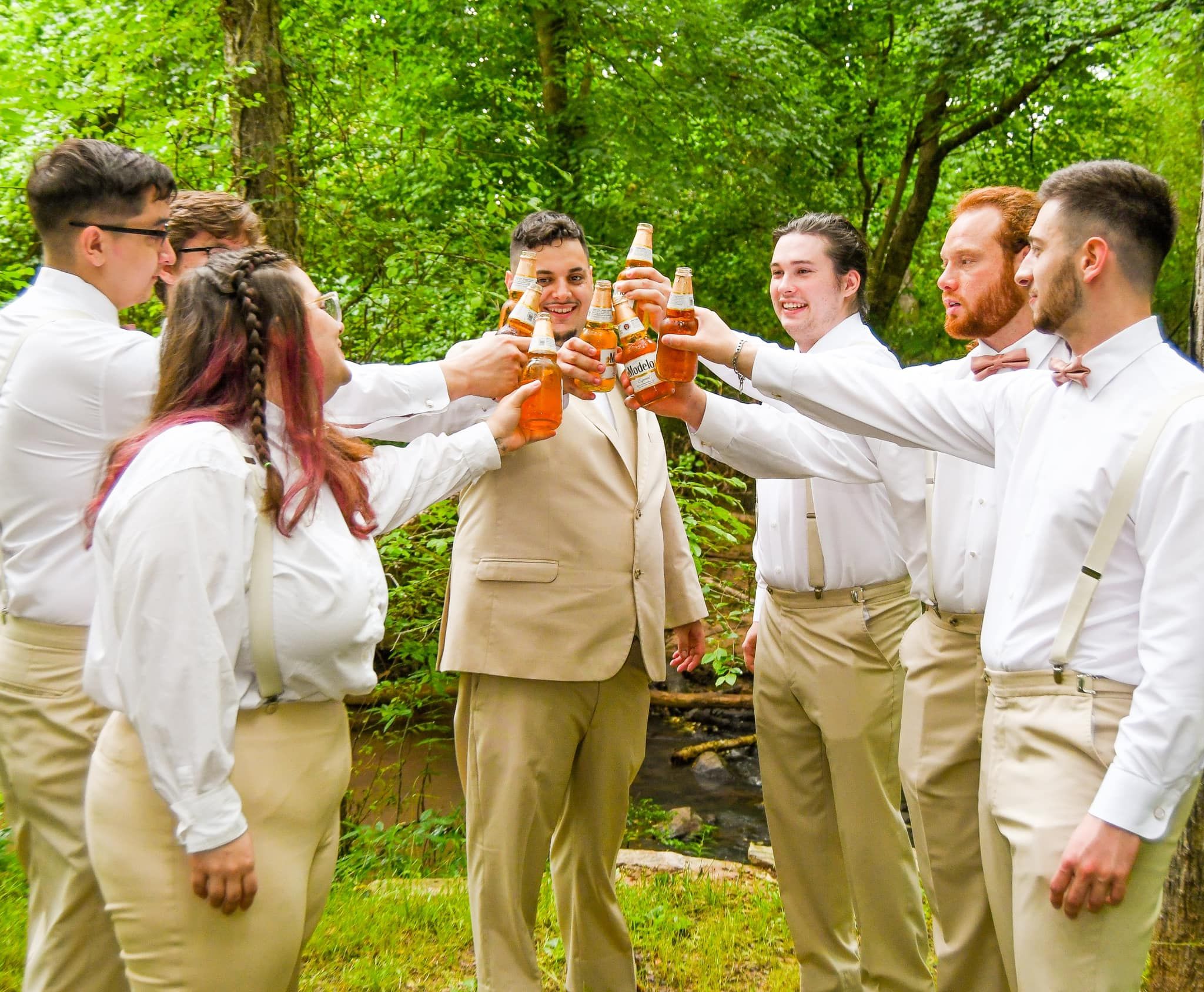 Wedding party in khaki and white attire toasting drinks together in a wooded outdoor setting.