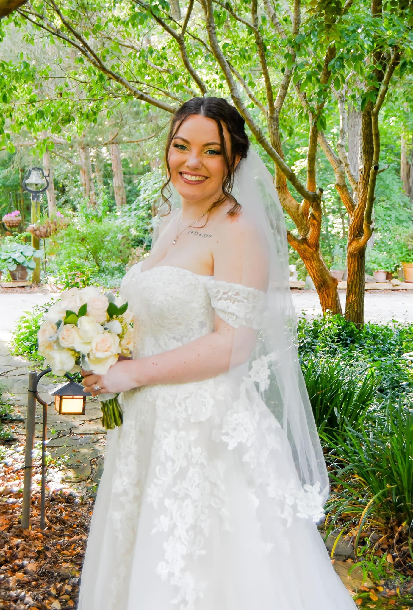 A bride in a white lace off-shoulder wedding gown holds a bouquet of white flowers in a garden setting.