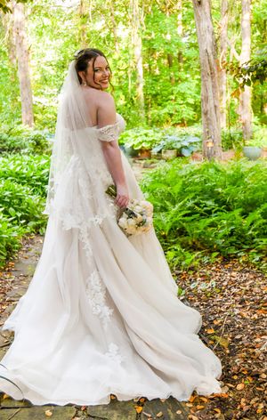 A bride in a flowing lace gown with long train poses in a wooded garden setting while looking over her shoulder.