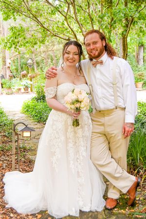 A wedding couple poses together in a lush garden setting wearing white and cream colored attire with bouquets.