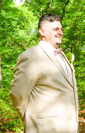 A person in a beige suit jacket stands in a lush green outdoor setting.