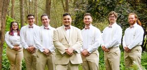 A wedding ceremony takes place under a white draped arch in a lush green forest setting.