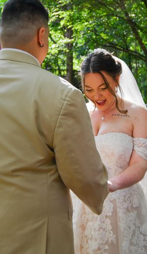 A bride in a lace wedding dress and groom in tan suit share a tender moment in a sunlit outdoor setting.
