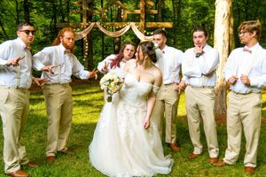 Wedding party poses playfully in line outdoors with groomsmen in beige suits and bridesmaids in white dresses.
