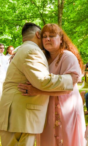 Two people embrace in an emotional moment during an outdoor ceremony surrounded by green trees.