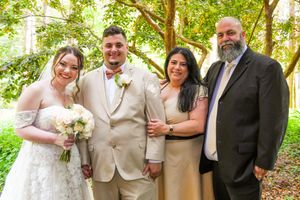 Wedding party poses outdoors under green trees in formal attire during summer celebration.