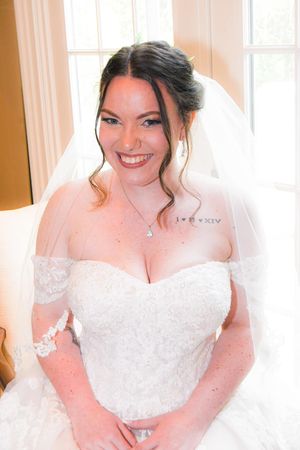 A bride in a white strapless lace wedding dress and veil smiles warmly while sitting in a sunlit room.