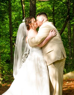 A romantic wedding couple shares tender kisses in a lush green forest setting during their summer ceremony.