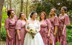 A row of bridesmaids in dusty rose dresses stand with the bride in a white gown outdoors in a garden setting.