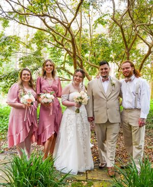 Wedding party in pink dresses and tan suits poses together outdoors under trees with greenery in the background.