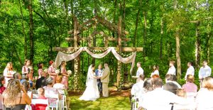 A panoramic view of an outdoor forest wedding ceremony with white chairs arranged in rows under tall green trees.