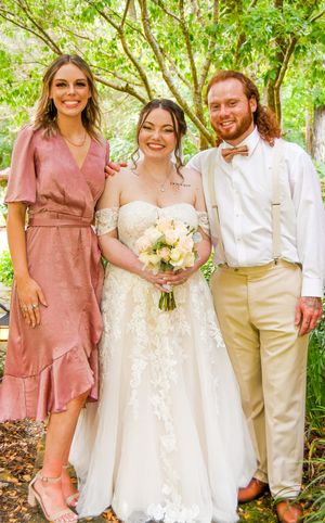 A wedding party poses outdoors in spring attire with pink and white color scheme against a leafy green backdrop.