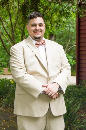 A wedding party member in a cream colored suit poses outdoors against a natural green background.
