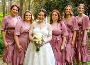 Wedding party poses outdoors in coordinated mauve dresses and white gown against natural forest backdrop.