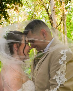 A romantic wedding couple shares tender kisses in a lush garden setting with sunlight filtering through the trees.