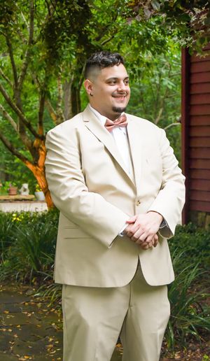 A person in a cream-colored suit poses outdoors against a lush green garden background.