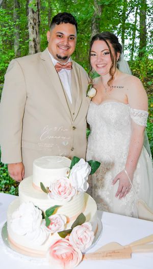 Newlyweds cut their elegant white wedding cake decorated with pink roses in an outdoor garden setting.