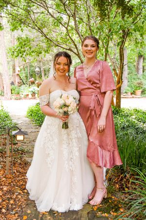 Two women pose in a garden setting, one wearing a white lace wedding gown and holding a bouquet, the other in a dusty rose dress.