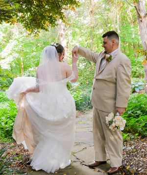 A couple in wedding attire twirls playfully on a garden path surrounded by lush green foliage.
