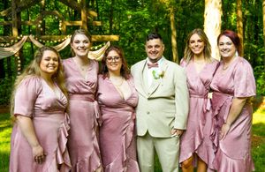 Wedding party in pink bridesmaid dresses and beige suits pose together in front of lush green foliage.