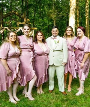 Wedding party wearing matching pink dresses and white shoes poses together in a lush green outdoor setting.