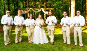 A wedding party in white dresses and khaki suits poses in a line outdoors against a green forest backdrop.