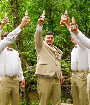 Groomsmen in tan suits raise drinks in celebratory toast outdoors.