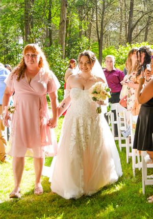 A wedding party in light pink dresses poses together on a sunny day in a garden setting with white chairs.