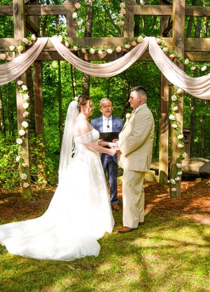 A romantic wedding ceremony takes place in a wooded outdoor venue decorated with flowing white drapes and string lights.