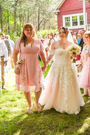 A wedding celebration takes place outside a red house with guests in pink dresses gathered on a sunny lawn.