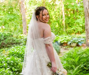A bride in a flowing white dress stands in a lush green garden setting during a summer wedding photoshoot.