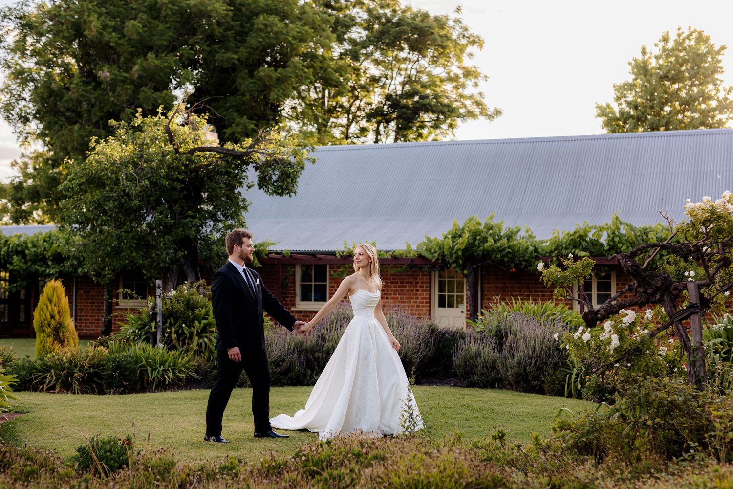 A couple stands together on a lush green lawn outside a rustic brick building at golden hour.