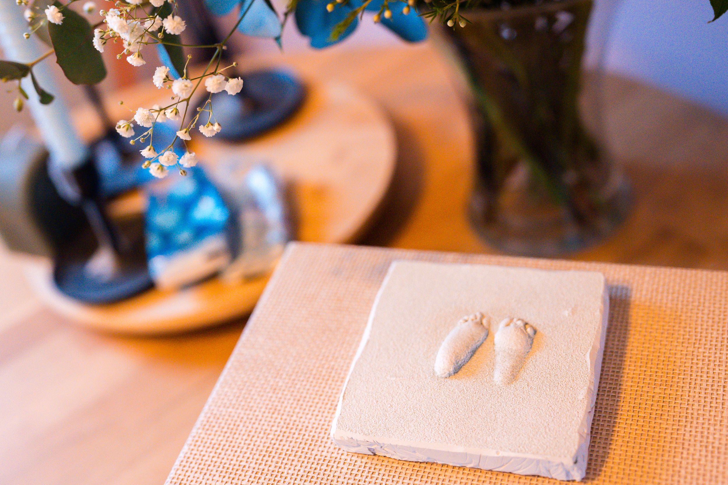 Small white baby footprint decorations rest on a pink textured surface near delicate floral arrangements.