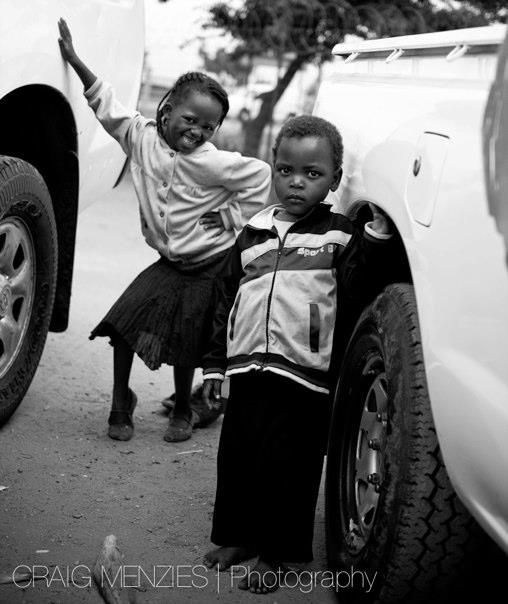 Black and white photo of two young African children smiling beside a white vehicle.