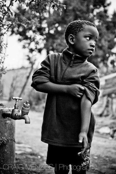 Black and white portrait of a young African boy standing near an outdoor water faucet.