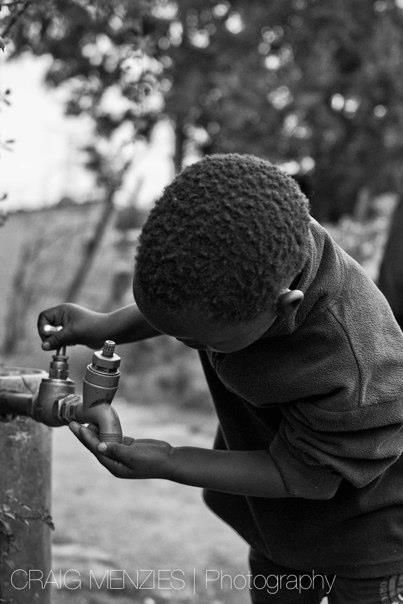 Black and white photo of a child drinking water from an outdoor metal tap.