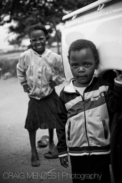 Black and white portrait of two young African children standing near a white vehicle.
