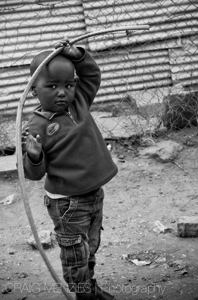 Young African toddler holding a hoop near a chain-link fence in black and white.
