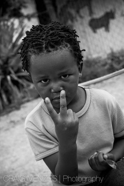 Young African child with dreadlocks pointing finger at camera in black and white portrait.