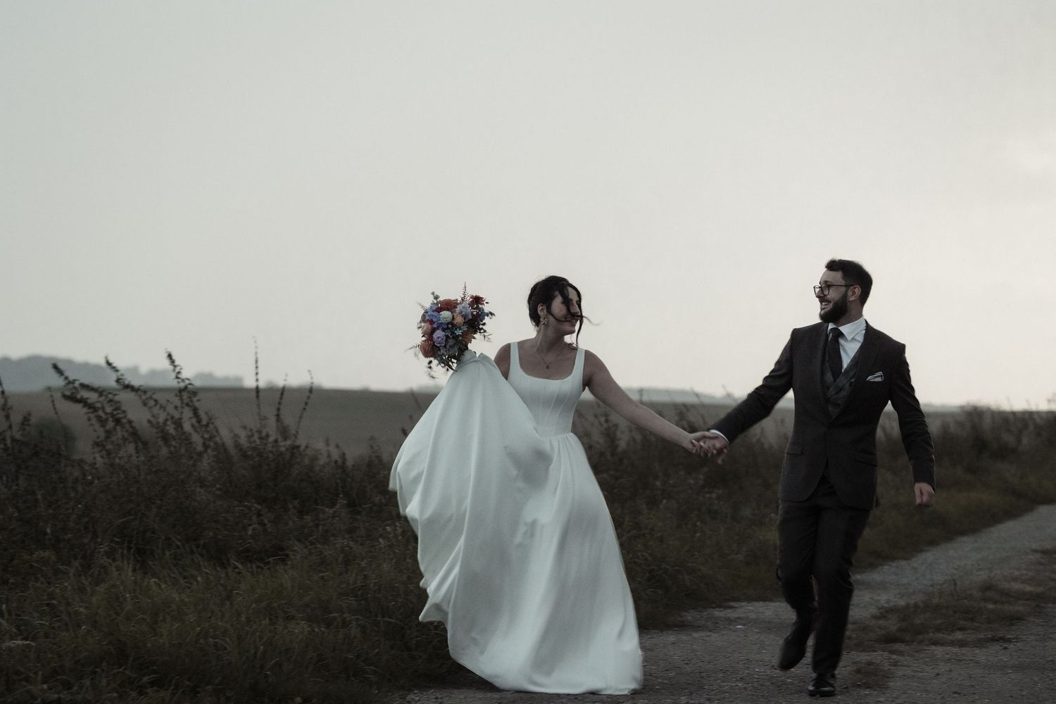 A couple in wedding attire runs playfully along a coastal path at sunset creating a romantic silhouette.