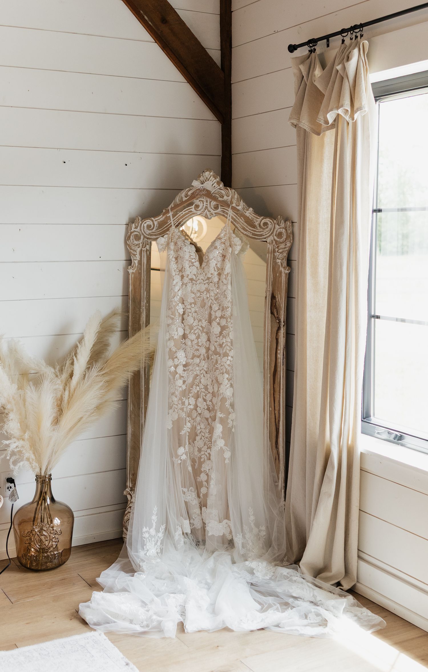 A romantic wedding dress displayed in a bright white room with natural light and pampas grass decor.