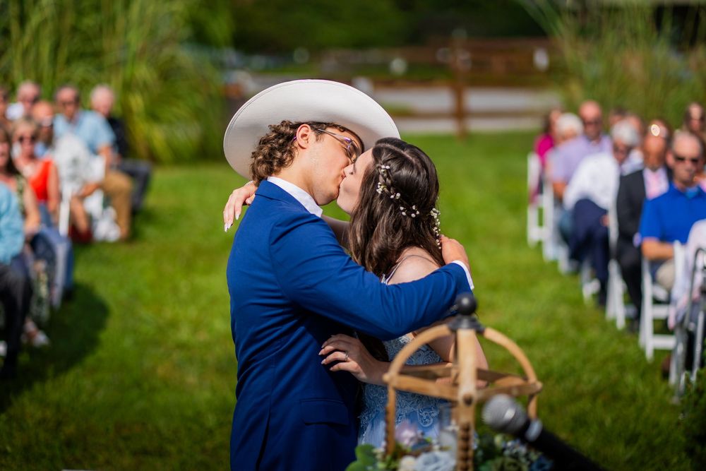 A couple in navy blue wedding attire share a romantic kiss during an outdoor ceremony with guests looking on.