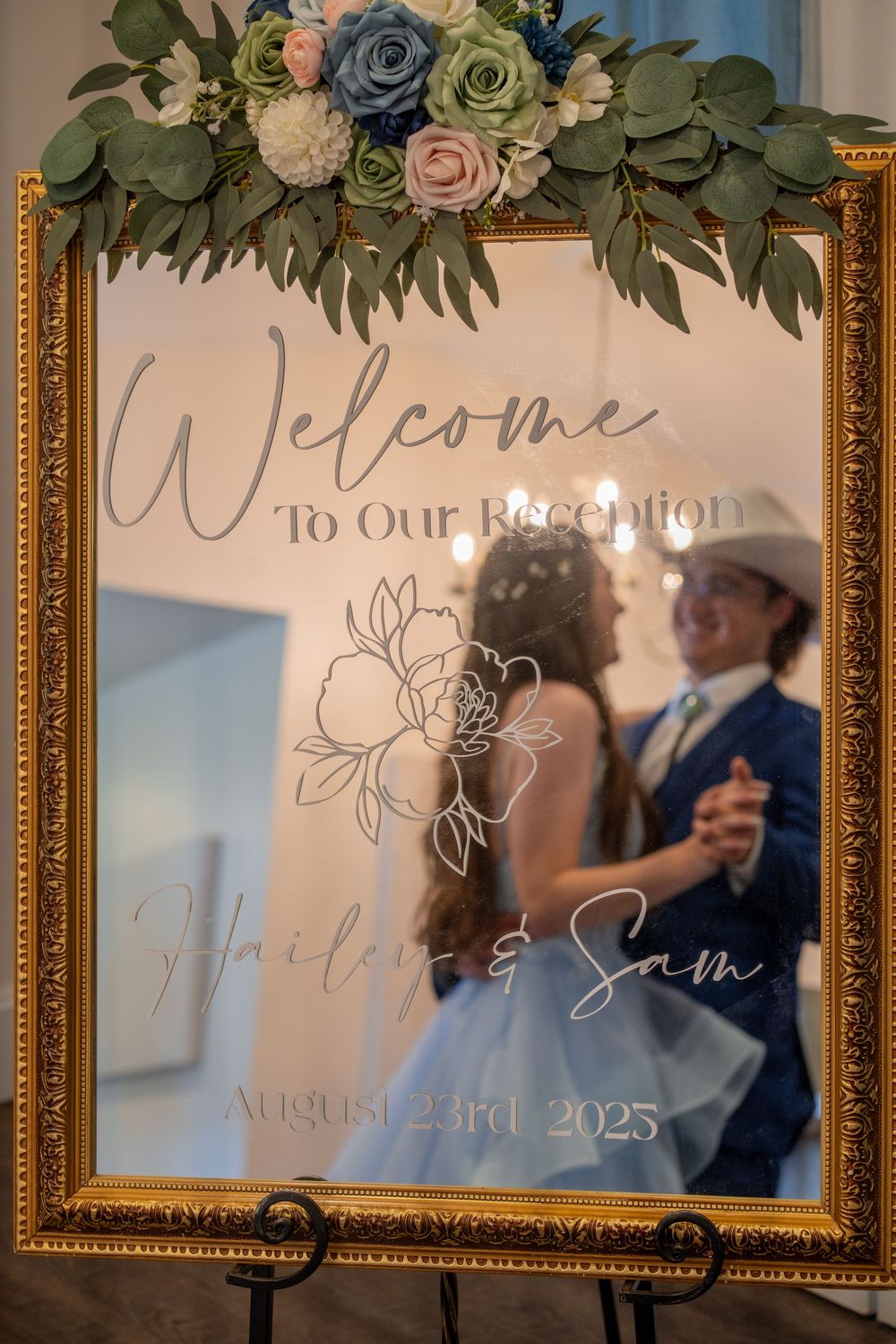 Elegant wedding reception welcome sign with floral decorations and gold frames displays event details.
